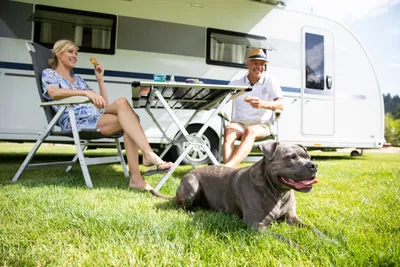 Alpsee Camping - Camperpaar mit Hund vor ihrem Wohnwagen in der Sonne