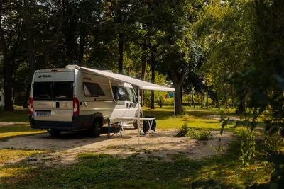Weißes Wohnmobil mit Markise und Campingstühlen am Stellplatz im Grünen, umgeben von Bäumen im Wald.