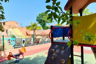 Spielbereich mit Rutsche und Kletterwand auf dem Campingplatz, im Hintergrund Pool, Liegen und Zäune.