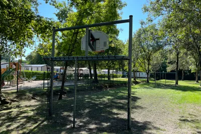 Spielplatz mit eingezäuntem Klettergerüst auf einem Campingplatz, umgeben von Bäumen und Rasenfläche.