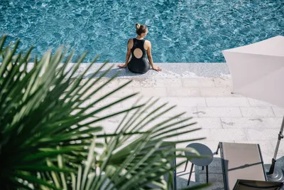 Frau sitzt am Poolrand auf einer Steintreppe, im Vordergrund Liegestuhl und Sonnenschirm, Palmenblätter im Bild.