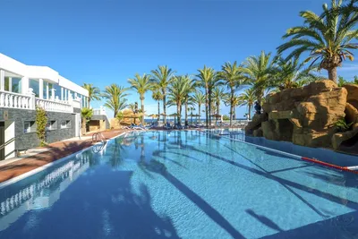Schwimmbecken eines Campingplatzes mit Palmen, Sonnenschirmen und Schattenwurf, im Hintergrund ein weißes Gebäude mit Balkon.