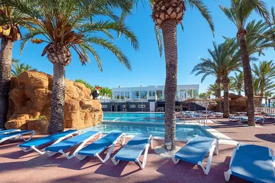 Poolbereich mit blauen Liegen, Palmen und Sonnenschirmen vor einem weißen Gebäude am Campingplatz.