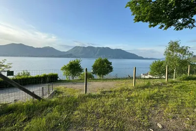 Campingplatz am See mit eingezäuntem Ufer, Grasfläche und Bäumen, im Hintergrund Berge und ruhiges Wasser.