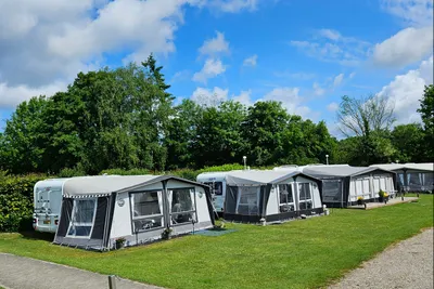 Reihe von Wohnwagen mit Vorzelten auf grünem Campingplatz, davor Rasen und im Hintergrund hohe Bäume und blauer Himmel.