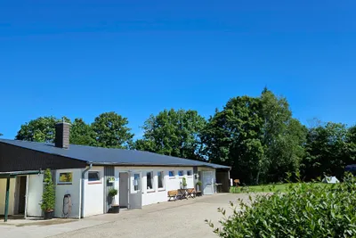 Campinggebäude mit Terrasse, Bank und Fahrradständer, umgeben von Bäumen, Blick in den blauen Himmel.