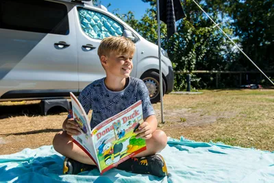 Camping De Kiekduun - Kind sitzt vor einem Standplatz und liest eine Zeitschrift
