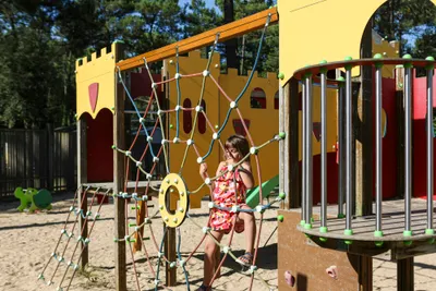 Spielplatz am Campingplatz mit Klettergerüst, bunten Netzen und einem Kind auf dem Sandboden.