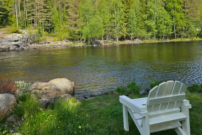 Weißer Campingstuhl am Seeufer mit Gras, Steinen und Blick auf den ruhigen Fluss sowie dichten Wald im Hintergrund.