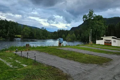 Uferpromenade am Fluss mit Kiesweg, Bäumen und einem kleinen Gebäude am Campingplatz, im Hintergrund bewaldete Hügel.
