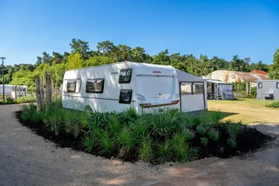 Weißer Wohnwagen mit Vorzelt und Fenstern, umgeben von Grasbeeten und Bäumen auf dem Campingplatz.
