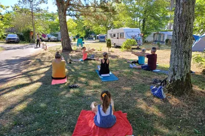 Campingplatz mit Wohnwagen und mehreren Personen beim Yoga auf Matten im Gras zwischen Bäumen.