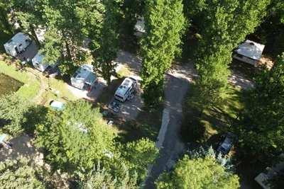 Luftaufnahme eines Campingplatzes mit Stellplätzen, Wohnwagen und Wohnmobilen, schmalem Weg und viel Grün zwischen Bäumen.