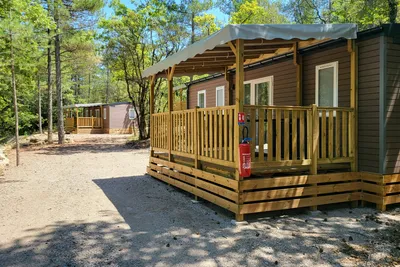 Holzbungalow mit Veranda und Geländer, daneben ein Feuerlöscher, gepflasterter Weg und Bäume im Hintergrund.