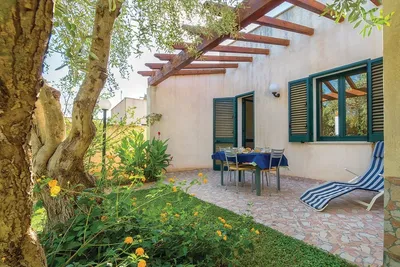 Terrasse am Campingplatz mit Tisch und Stühlen, Sonnenschirm, Liegestuhl und Blumenbeeten vor einer weißen Hauswand.