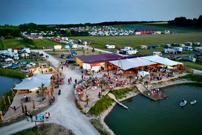 Luftaufnahme eines Campingplatzes mit Seezugang, Restaurant mit Terrasse, Booten und Wohnwagen entlang eines Weges.