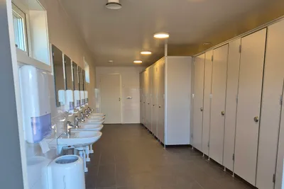 Reihe weißer Toiletten und Waschbecken in einem hellen Sanitärraum mit Kabinen und Fliesenboden.