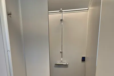 Barrierefreie Duschkabine mit Duschkopf, Haltegriffen und grauen Fliesen, daneben eine Ablage und eine Tür.